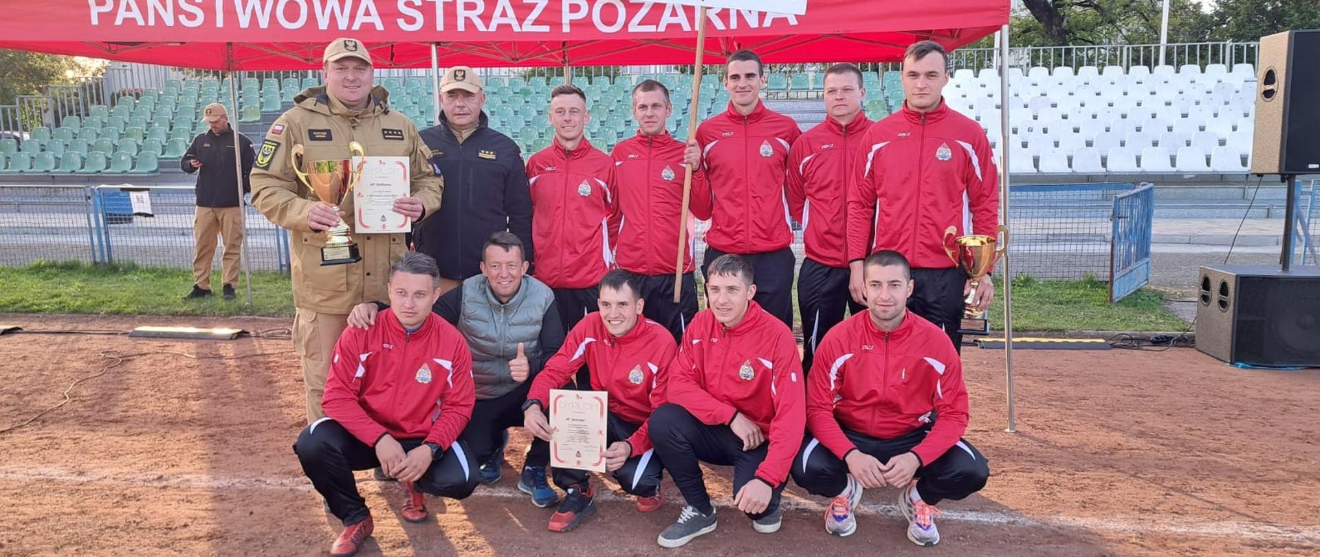 Zdjęcie grupowe strażaków w strojach sportowych koloru czerwonego wraz z Komendantem Powiatowym PSP oraz Dowódcami Jednostki Ratowniczo-Gaśniczej.