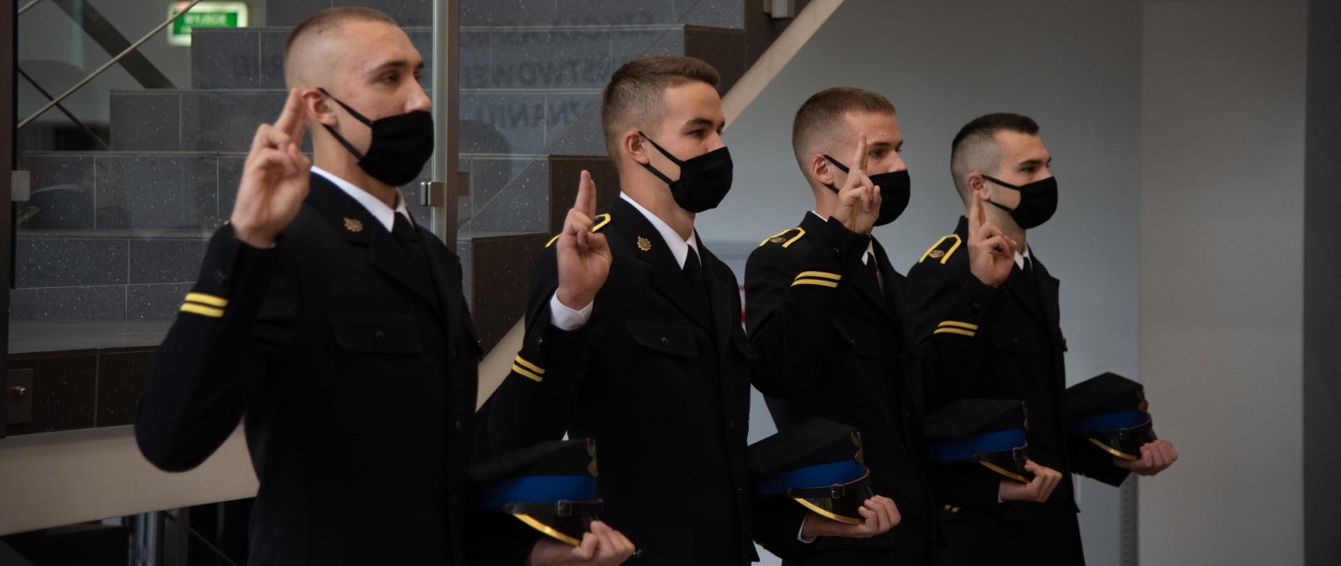 Trzech młodych strażaków w mundurach wyjściowych w lewej ręce trzymają rogatywki, Prawe dłonie są uniesione z dwoma palcami do góry, jak do ślubowania