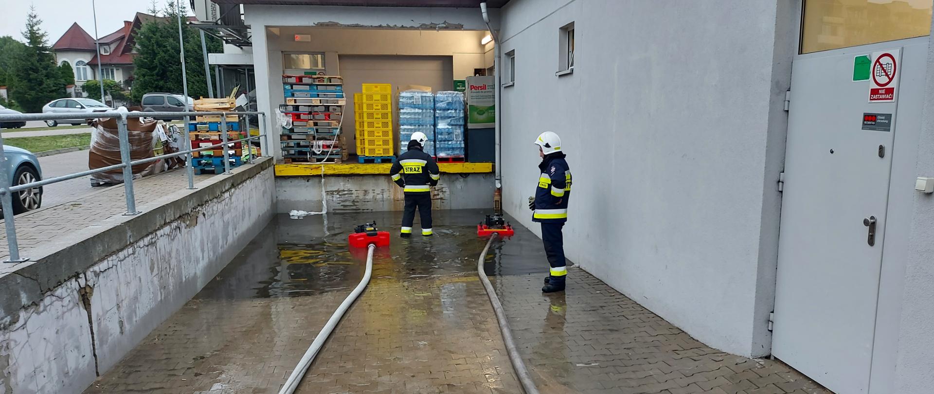 Strażacy OSP nadzorujący motopompy pływające wypompowują wodę z podjazdu rampy załadunkowej w sklepie wielkopowierzchniowym.