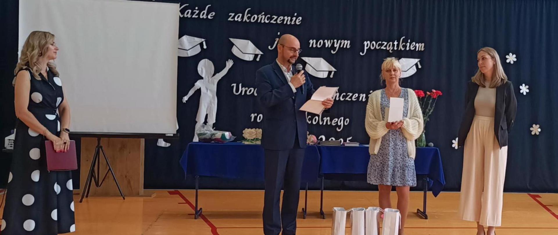 na zdjęciu trzy osoby, wszyscy stoją, pierwsza z lewej strony stoi kobieta ubrana w długą granatową w duże, białe grochy sukienkę obok mężczyzna ubrany w ciemny garnitur przemawia z mikrofonem w ręku, obok niego stoi kobieta ubrana w jasną sukienkę do kolan i biały sweterek, kolejna osoba to kobieta ubrana w jasne spodnie i bluzkę oraz czarny żakiet, 