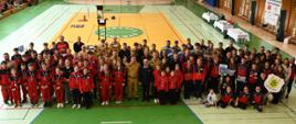 Zbiorowa fotografia dużej grupy dzieci, młodzieży w dresach i kaskach oraz strażaków w mundurach zebranych na parkiecie hali sportowej po zakończeniu zawodów pożarniczych.
