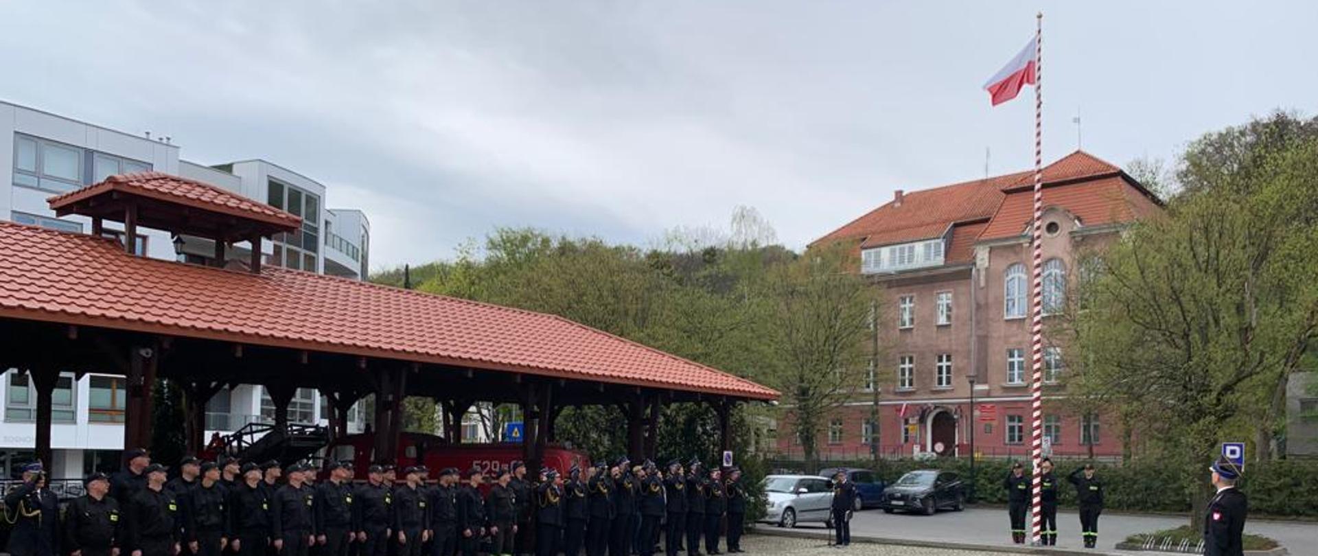 Strażacy stoją na zbiórce podczas podnoszenia flagi na maszt.