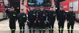 Strażacy w szeregu stoją na tle samochodów strażackich
