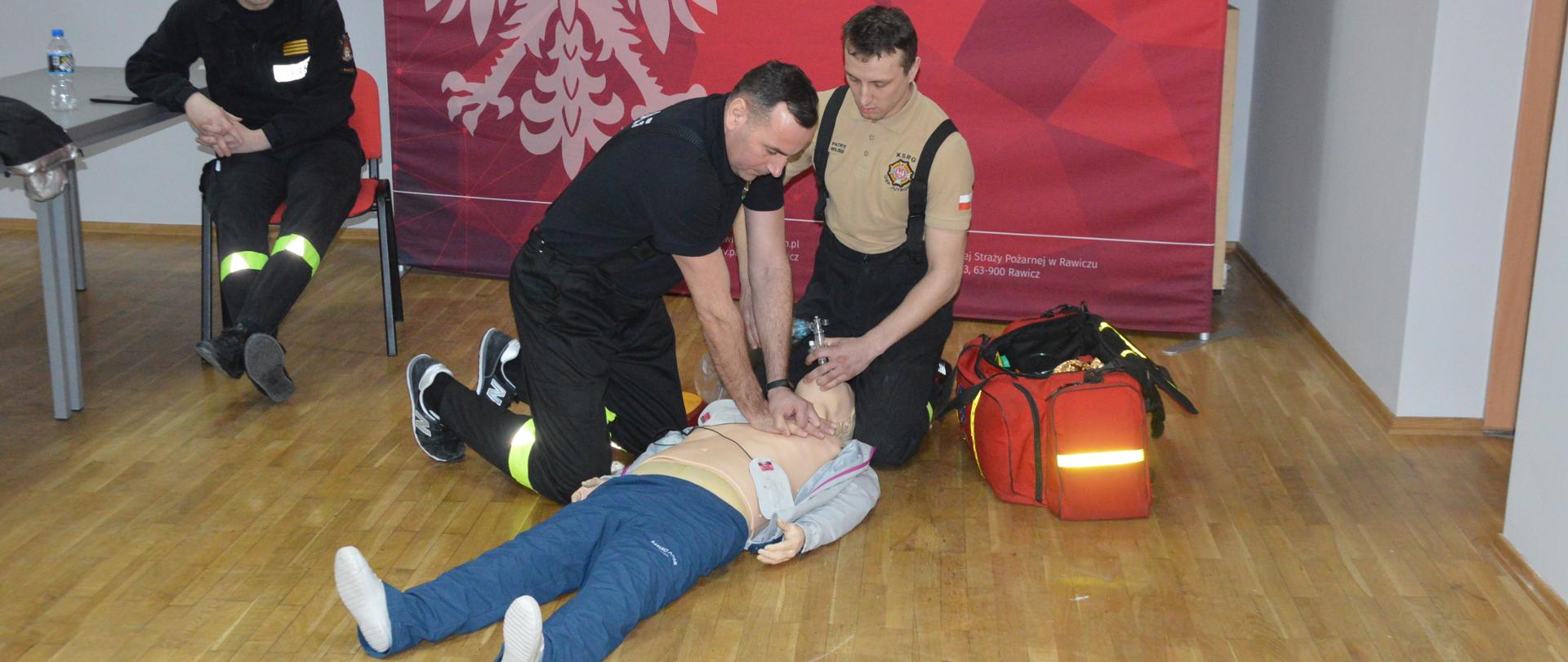 Egzamin recertyfikacyjny. Świetlica Komendy Powiatowej PSP w Rawiczu. Na tle ścianki promującej komendę dwóch egzaminowanych strażaków ratowników OSP udziela pomocy "poszkodowanemu" manekinowi. Jeden z nich klęcząc z bok wykonuje zewnętrzny masaż serca , drugi, znajdujący sią za głową - przy użyciu worka samorozprężalnego prowadzi sztuczny oddech. Z prawej strony stoi torba medyczna. Do ciała poszkodowanego przyklejone są elektrody AED . Przy stole siedzi członek komisji egzaminacyjnej.