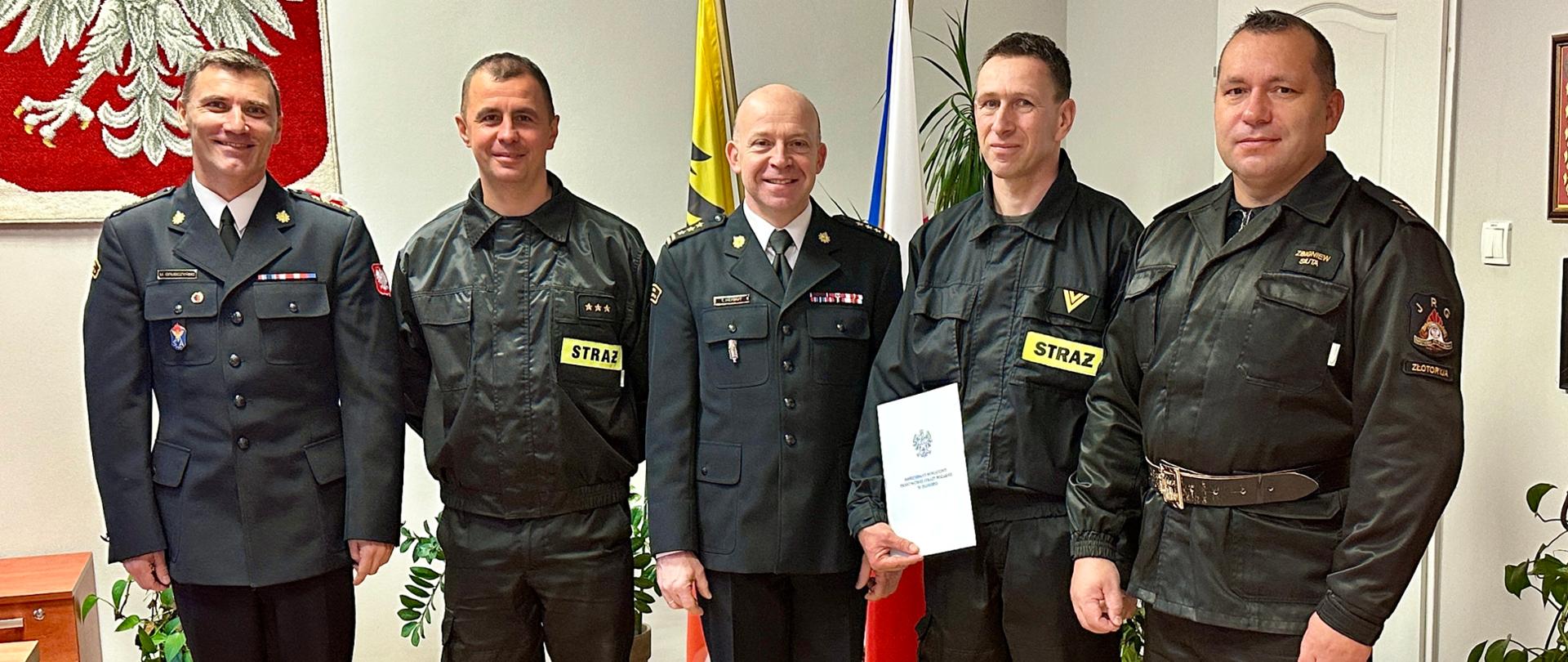 pięciu strażaków w gabinecie komendanta, w tle godło państwowe oraz flaga