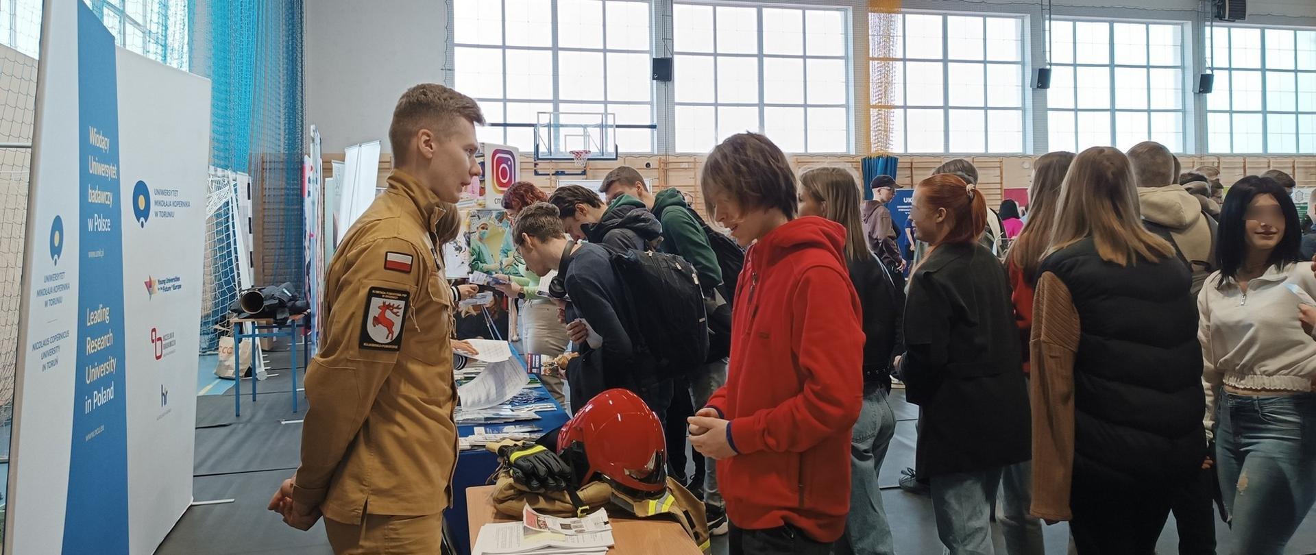 Zdjęcie przedstawia uczestników Targów Edukacji i Pracy przed stoiskiem Komendy Powiatowej PSP w Brodnicy. Po lewo ubrany w mundur służbowy przedstawiciel Komendy PSP w Brodnicy.
