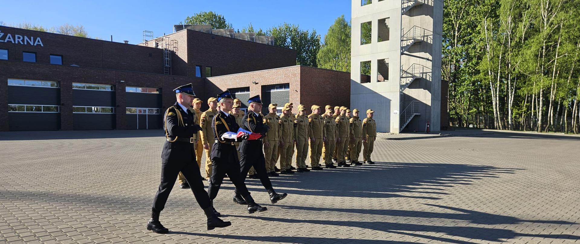 Na placu przed budynkiem straży pożarnej trzech strażaków w galowych mundurach niesie złożoną flagę, maszerując przed ustawioną w szeregu grupą strażaków w beżowych mundurach. W tle widoczna jest wieża ćwiczeniowa i drzewa, a długie cienie wskazują na niskie położenie słońca.