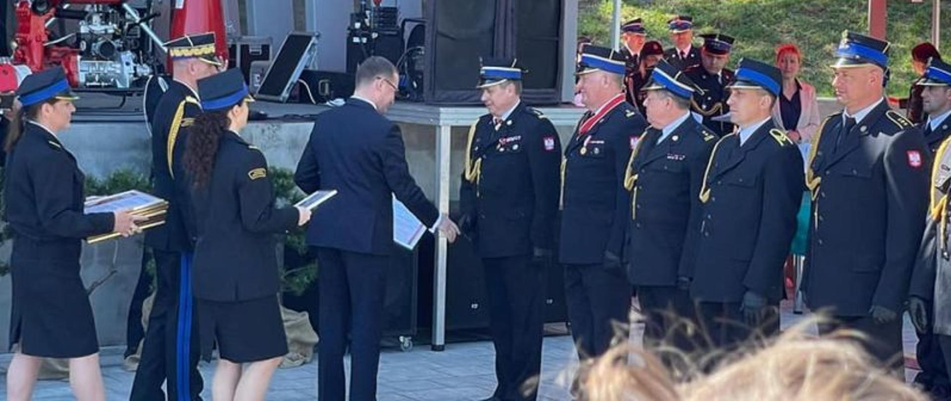 Zdjęcie wykonane w dzień. W tle scena amfiteatru. Na pierwszym planie strażacy w mundurach galowych przyjmujący odznaczenia. Dzień pogodny, słoneczny.