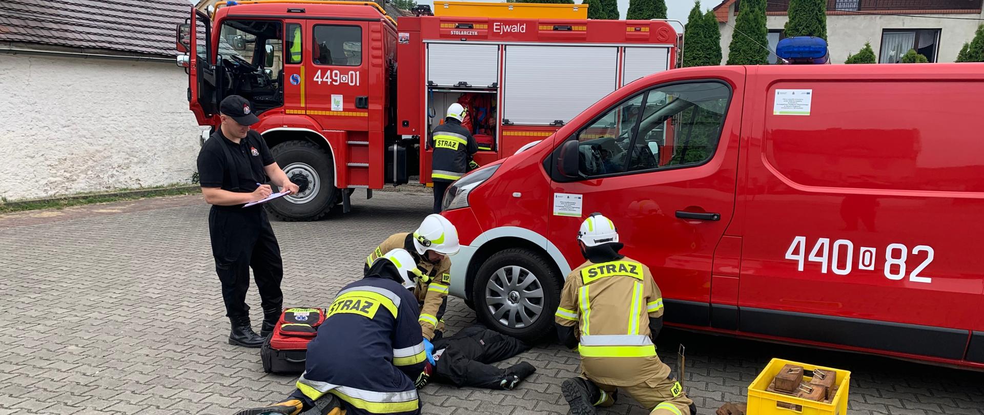 W tle samochód pożarniczy OSP. Na pierwszym planie dwóch ratowników OSP udziela pomocy poszkodowanemu (manekin) przyciśniętemu kołem samochodu PSP (furgon) Trzeci ratownik stabilizuje pojazd przy pomocy klinów. Obok stoi funkcjonariusz PSP z podkładką na dokumenty oceniający inspekcję. Czwarty ratownik OSP stoi przy samochodzie OSP wyciągając potrzebny w akcji sprzęt. Strażacy w Ubraniach specjalnych i białych hełmach.
