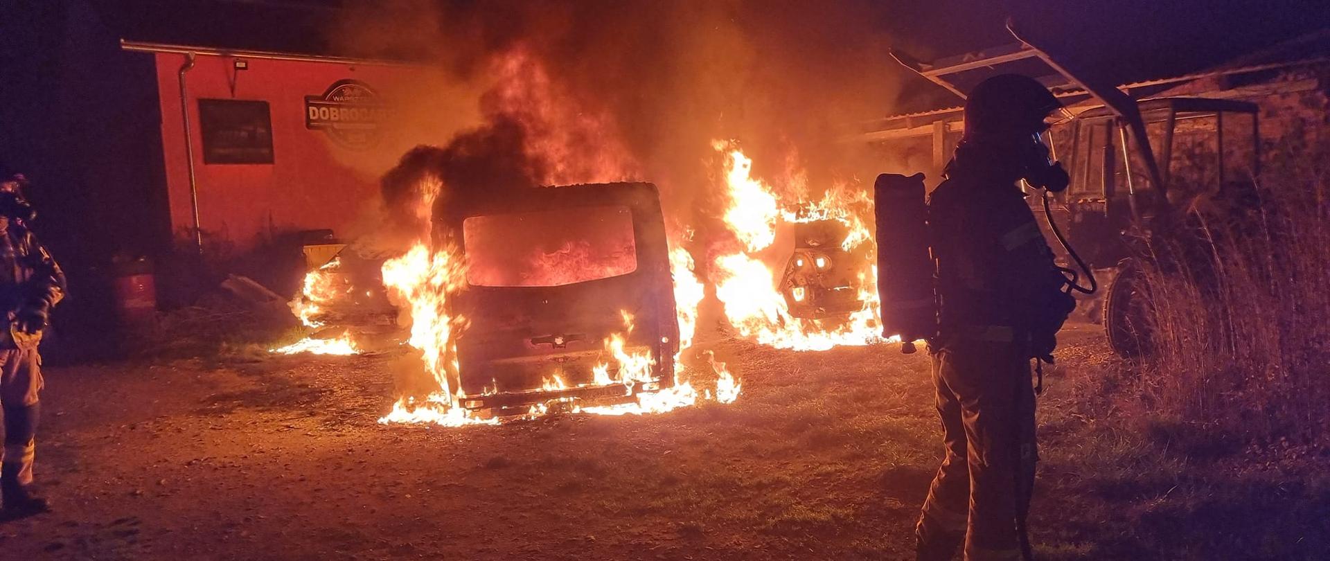 Widać pożar dwóch samochodów, jest ogień i ciemno, po lewej i prawej jest strażak