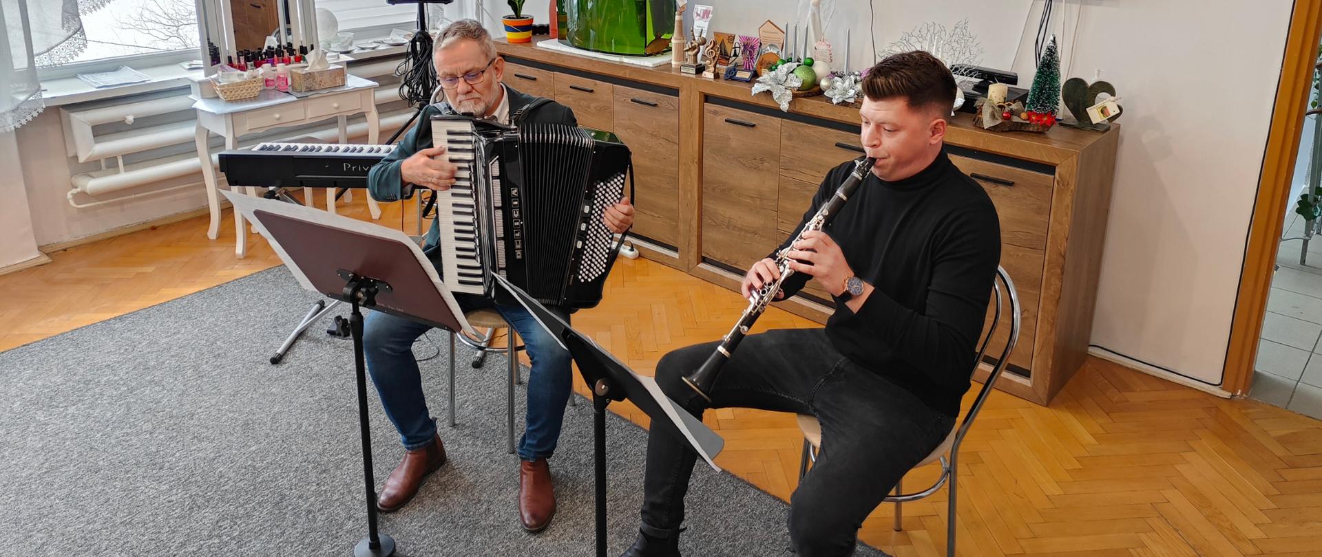 Na zdjęciu widać dwóch mężczyzn. Obaj siedzą na jasnych krzesłach. Przed nimi stoją pulpity na nuty. Mężczyzna siedzący z prawej strony jest ubrany na czarno i gra na czarnym klarnecie. Mężczyzna siedzący z lewej strony jest ubrany w niebieskie spodnie, białą koszulę, ciemną marynarkę i gra na czarnym akordeonie. W tle widać brązową komodę z dekoracją świąteczną, telewizor zawieszony na jasnej ścianie, czarny instrument klawiszowy na statywie oraz kolumnę głośnika na statywie.