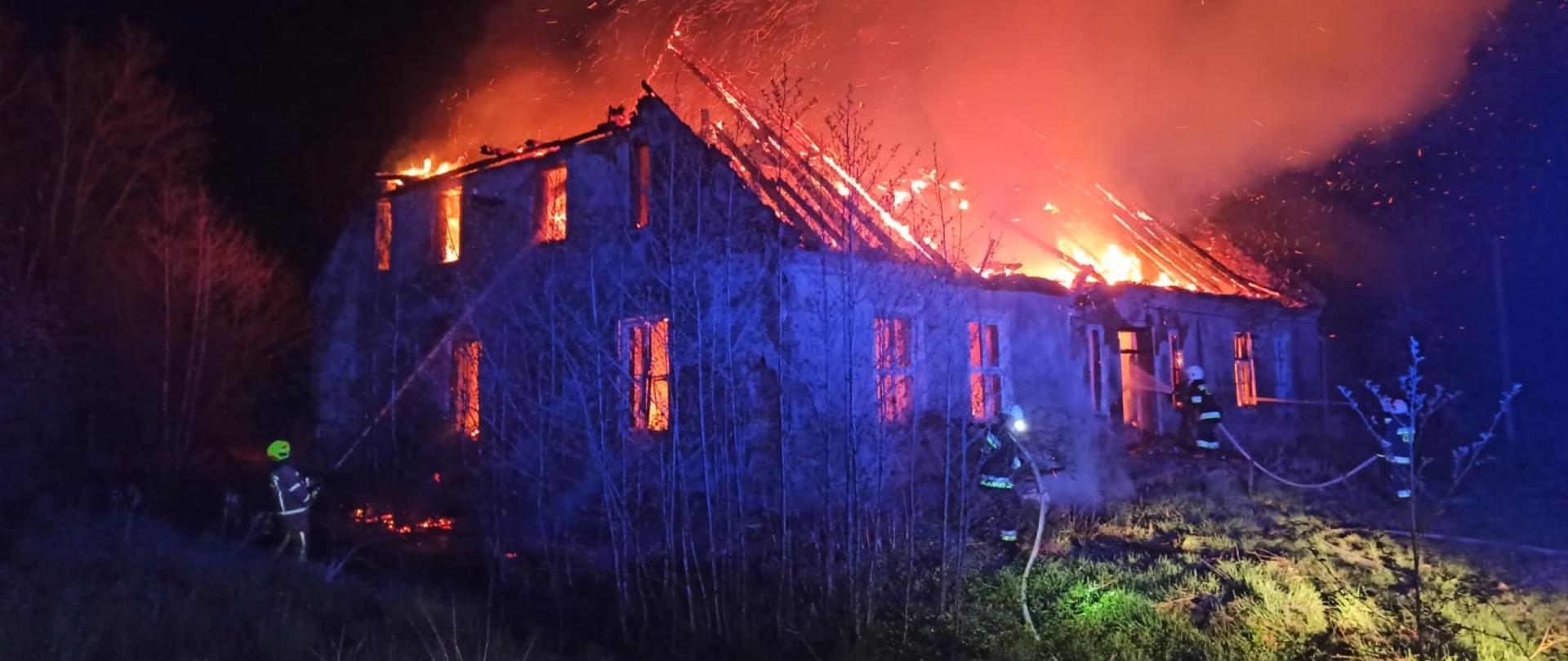 Zdjęcie zrobione nocą. Dach budynku mieszkalnego cały w ogniu. Strażacy gaszą pożar.