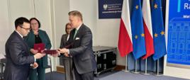 Prof. Robert Gawłowski odbiera akt powołania z rąk min. Jakuba Stefaniaka, sekretarza stanu, Zastępcy Szefa Kancelarii Prezesa Rady Ministrów; w tle flagi Polski i Unii Europejskiej