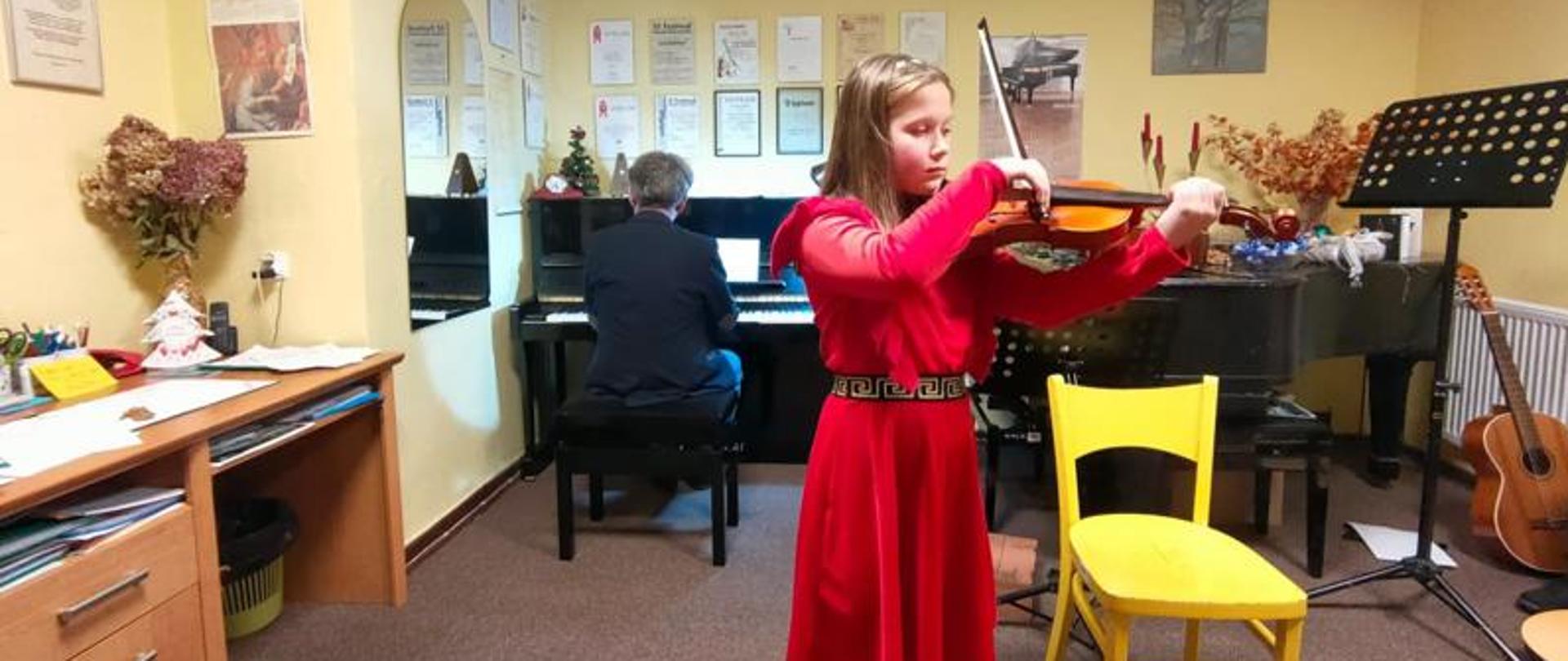 Zdjęcie kolorowe, dziewczynka w czerwonej sukience ze skrzypcami w rękach. Za dziewczynką przy pianinie siedzi akompaniator.