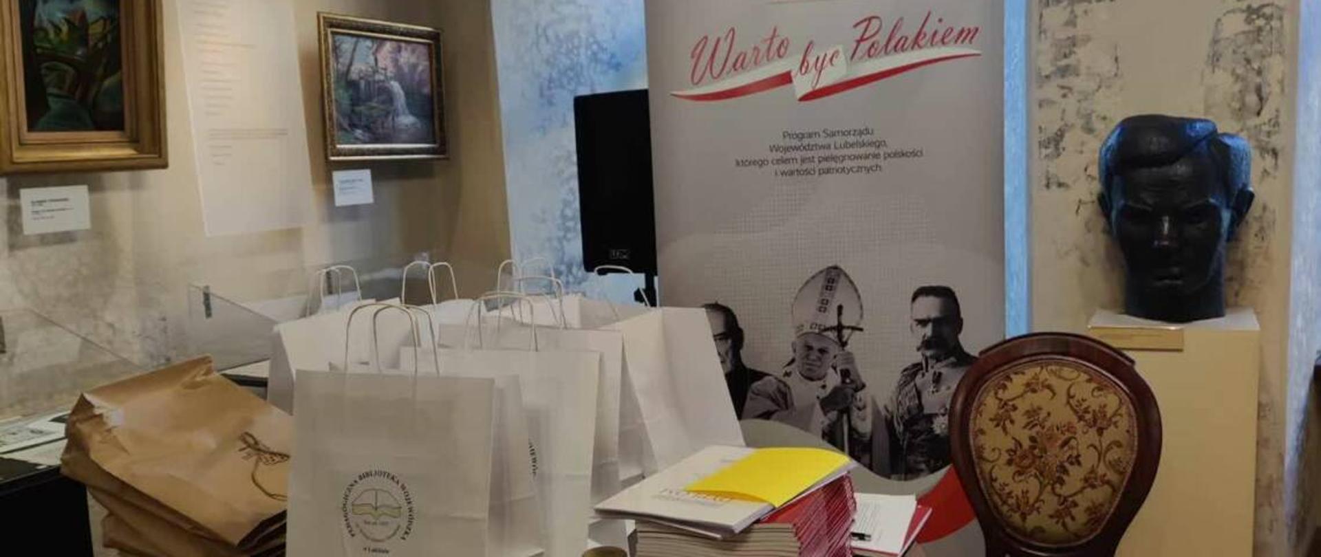 W sali Muzeum Józefa Czechowicza w Lublinie na okrągłym, stylowym stole znajdują się papierowe, szare i białe torby z prezentami oraz bordowe katalogi ułożone w stos. Po prawej strone stoi stylowe, tapicerowane krzesło. Za stołem znajduje się bilbord, na którego szarym tle są: logo Samorząd Województwa Lubelskiego - herb (biegnący biały jeleń na czerwonym tle), napisy: "Warto być Polakiem", "Program Samorząd Województwa Lubelskiego, którego celem jest pielęgnowanie polskości i wartości patriotycznych" oraz portrety trzech Polaków: Stefan Wyszyński, Jan Paweł II, Józef Klemens Piłsudski. Obok na kubiku stoi rzeźba - głowa. Na ścianach wiszą obrazy w złotych ramach.