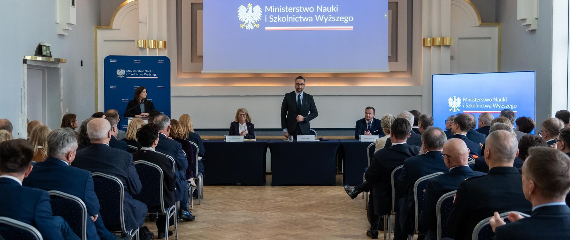 Widok od tyłu na salę, w rzędach krzeseł siedzą ludzie, przed nimi za czarnym stolikiem siedzi kobieta w czarnym ubraniu i mężczyzna w garniturze, między nimi stoi minister Kulasek, za nim na wielkim wiszącym pod sufitem ekranie napis Ministerstwo Nauki i Szkolnictwa Wyższego. 