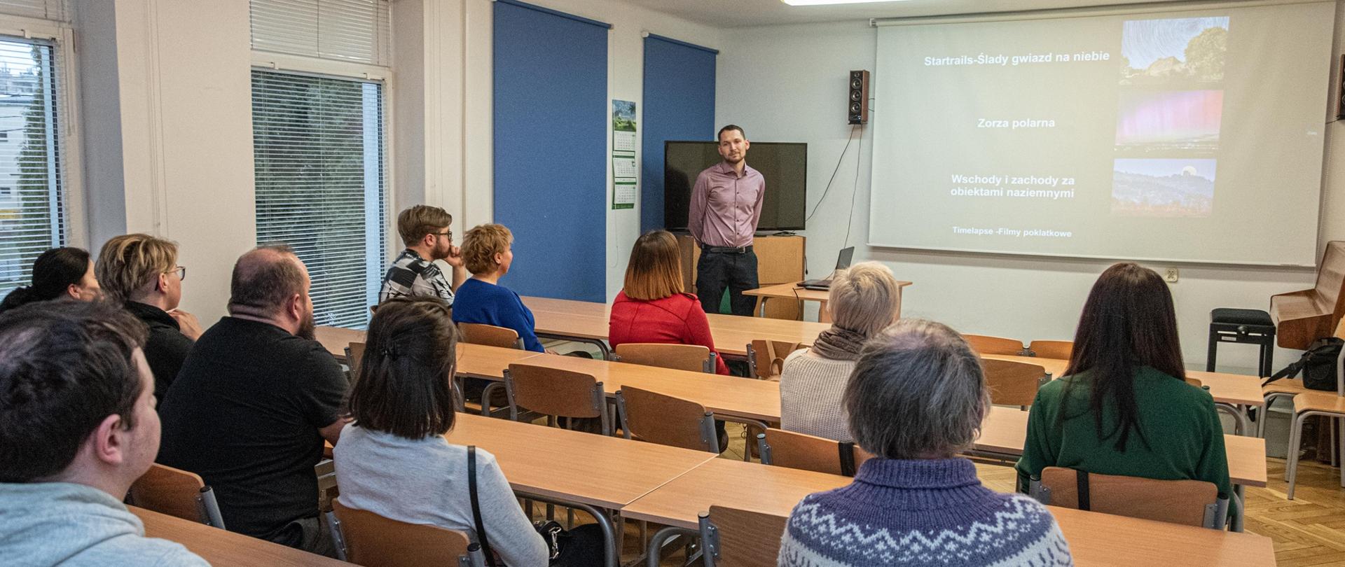 Zdjęcie przedstawia publiczność zgromadzoną w sali podczas prelekcji autora wystawy zatytułowanej Chwile zatrzymane w kadrze. Publiczność siedzi tyłem przy ławkach. Prowadzący prelekcję stoi przodem. Na ścianie wyświetlane są zdjęcia. 