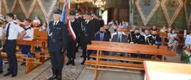 Na zdjęciu widoczny pododdział wraz z pocztem sztandarowym oraz siedzący zaproszeni goście w kościele podczas uroczystości poświęcenie i przekazani samochodu pożarniczego. W tle na ścianach kościoła widoczne polichromie na zielonym tle.