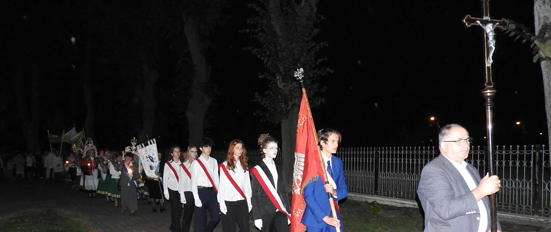 Nocna procesja religijna idąca alejką przy kościele. Na czele pochodu mężczyzna niesie krzyż, za nim młodzież w białych koszulach z czerwonymi szarfami i sztandarem. Dalej widać osoby w strojach ludowych niosące chorągwie i świece. W tle widać drzewa i ogrodzenie. Na dole zdjęcia widoczna jest data: 18.09.2025, godzina 19:27.