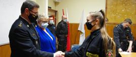 Widok z boku. Na pierwszym planie komendant powiatowy PSP w umundurowaniu podaje rękę druhnie OSP. Obok na drugim planie stoi wicestarosta wadowicka w niebieskim ubraniu cywilnym. W tle widocznych trzech strażaków PSP i OSP w umundurowaniu. Wszystkie osoby w maseczkach ochronnych. 