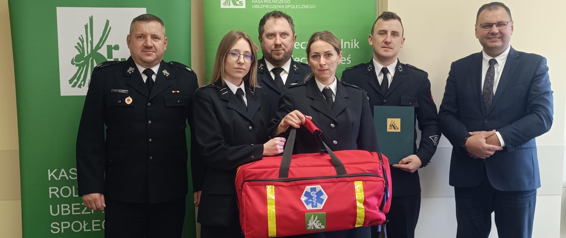 Grupa osób pozuje do zdjęcia w pomieszczeniu. Za nimi stoją dwa banery z logiem KRUS. W pierwszym rzędzie stoją dwie kobiety, druhny OSP. Za nimi stoi trzech strażaków oraz pierwszy od prawej mężczyzna w garniturze 