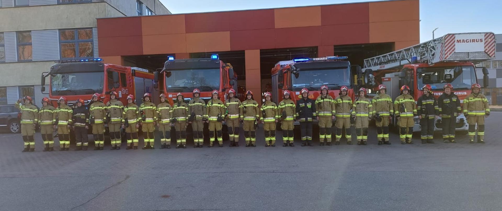 Na tle samochodów pożarniczych wystawionych z garażu stoją strażacy, dowódca salutuje