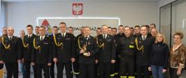 Koledzy emeryta, strażacy oraz Kierownictwo KP PSP w Radomsku w umundurowaniu. Emeryt trzyma pamiątkową statuetkę. Zdjęcie grupowe.
