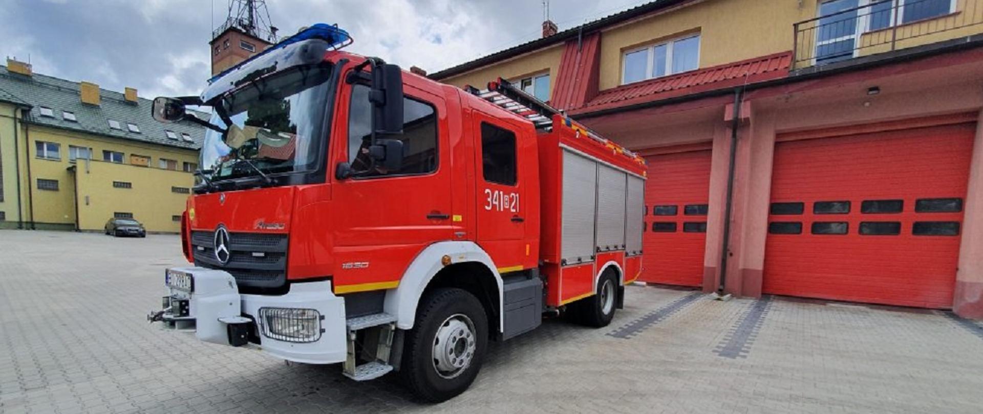 Samochód średni ratowniczo-gaśniczy z modułem technicznym w kolorze czerwony z białymi zderzakami. W drugim planie zdjęcia dwa budynki. Pierwszy budynek Jednostki Ratowniczo Gaśniczej numer 1 z bramami garażowymi koloru czerwonego. Drugi budynek w kolorze żółtym Stacji Ratownictwa Medycznego.
