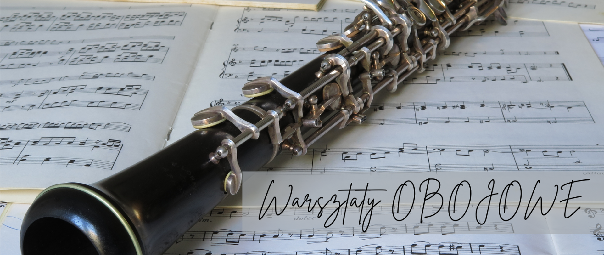 Na zdjęciu widać obój leżący na otwartych nutach. Instrument jest czarny z metalowymi klapami, a jego detale są dobrze widoczne. W tle rozłożone są kartki z zapisem nutowym. W prawym dolnym rogu znajduje się napis „Warsztaty OBOJOWE” zapisany elegancką, ozdobną czcionką.