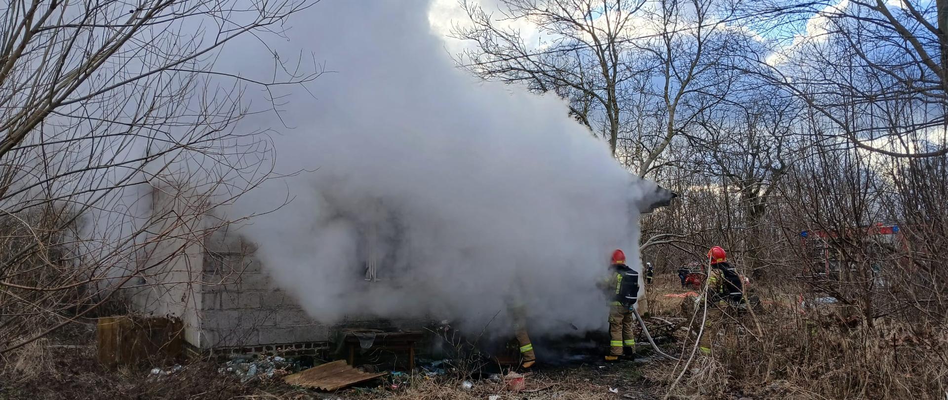 Zdjęcie przedstawia akcję gaśniczą przy niewielkim, parterowym budynku murowanym z jasnych bloczków. Obiekt jest niemal całkowicie przesłonięty gęstymi kłębami białego i jasnoszarego dymu, który wydobywa się z wnętrza i unosi wysoko do góry. Na pierwszym planie, po prawej stronie, widocznych jest dwóch strażaków w pełnym umundurowaniu bojowym i czerwonych hełmach, którzy kierują strumień wody w stronę źródła ognia. Teren wokół jest zaniedbany, porośnięty suchą trawą, krzewami i pozbawionymi liści drzewami; widać również rozrzucone śmieci. W tle, po prawej stronie, majaczy czerwony wóz strażacki. Niebo jest częściowo zachmurzone z przebijającym się światłem dziennym.