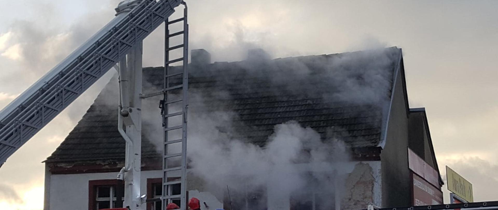 Dwa samochody strażackie koloru czerwonego jeden podnośnik i średni ratowniczo-gaśniczy, ustawione na ulicy Lipowej przy budynku z którego wydobywa się dym. 