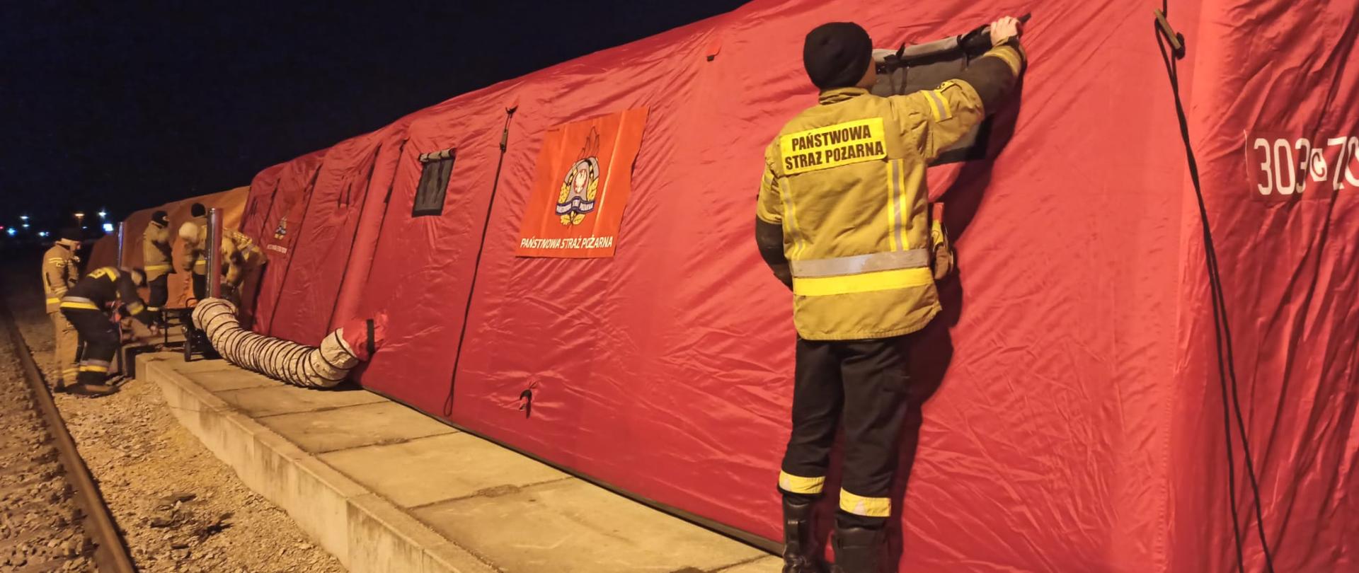 Strażak w żółtym ubraniu specjalnym stojący tyłem do osoby robiącej zdjęcie, strażak poprawia okienko w namiocie pneumatycznym koloru czerwonego. 