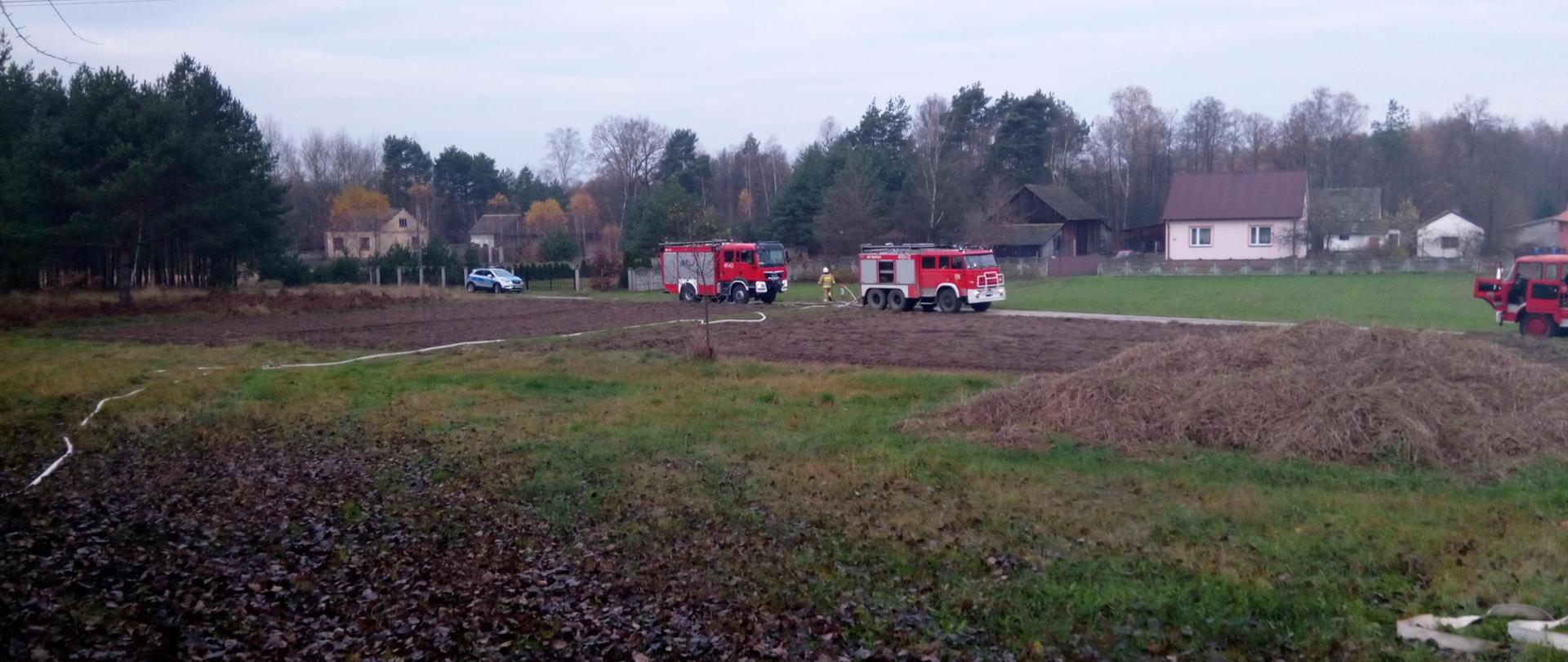 Widok z oddali na dwa samochody gaśnicze stojące na drodze od których poprowadzona jest linia gaśnicza, na drodze radiowóz Policji, w tle zabudowania w miejscowości.
