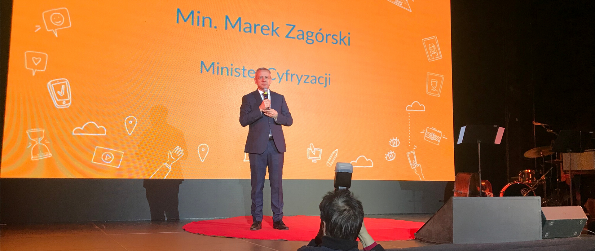 Minister Marek Zagórski na scenie z mikrofonem. Za nim ścianka z napisanymi (jego imieniem i nazwiskiem), funkcją oraz obrazkami - symbolami Internetu, np. chmura, otwarty laptop, wifi. Przed sceną fotograf z aparatem..