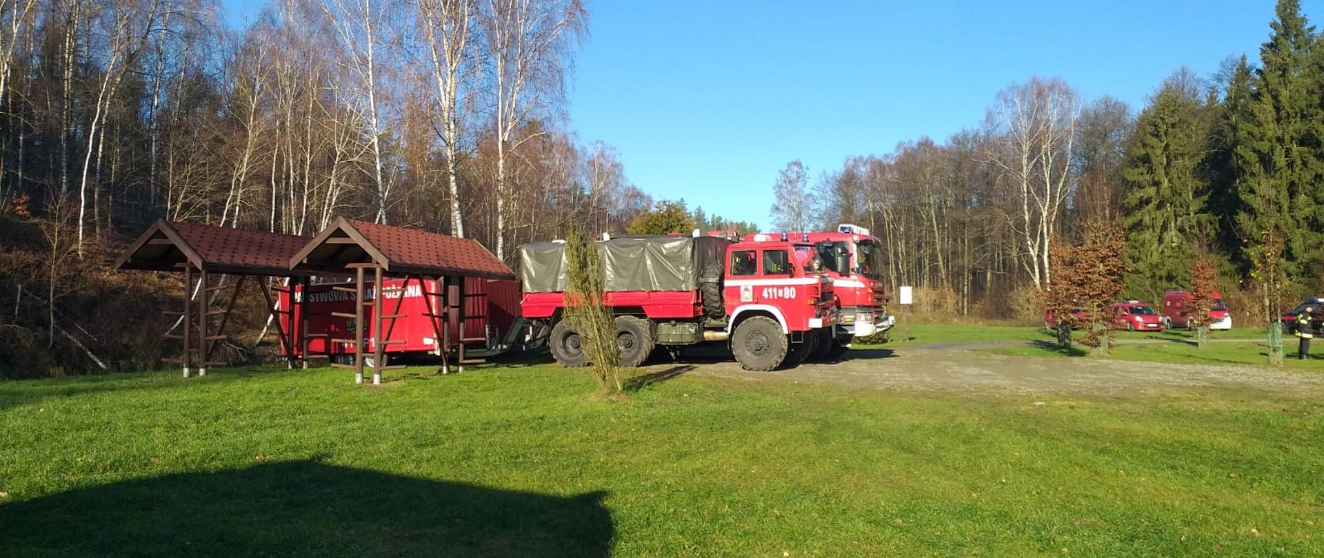 Na polanie w lesie stoją czerwone samochody strażackie. W tle las. Słoneczna jesienna pogoda.