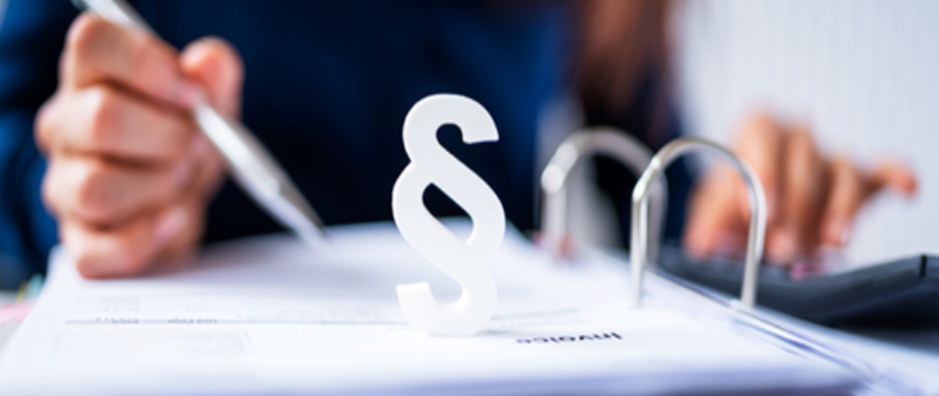 Na pierwszym planie symbol znak paragrafu, w tle w nieostrości segregator i ręka z długopisem