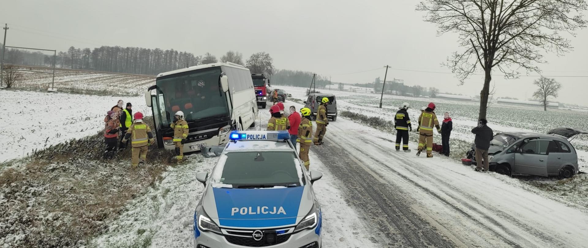 Na pierwszym planie stoi radiowóz policyjny. Po lewej stronie w przydrożnym rowie stoi autobus po kolizji z samochodem osobowym, który stoi po przeciwległej stronie jezdni w rowie. Kilku strażaków i poszkodowanych stoi obok aut.