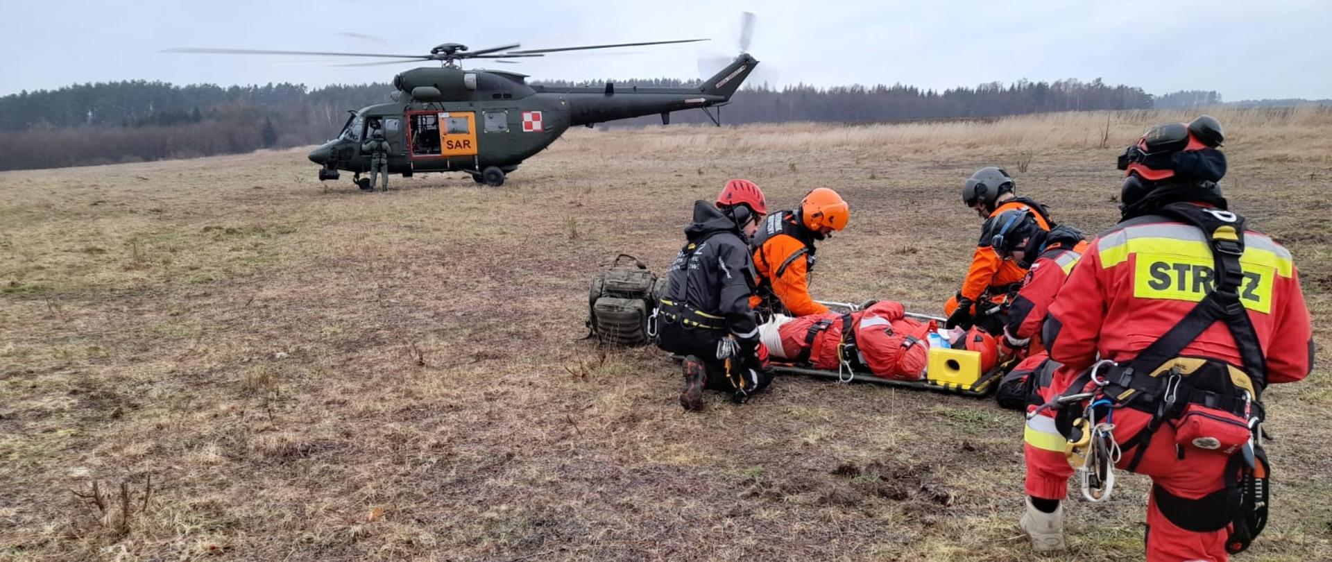 Zdjęcie przedstawia śmigłowiec oraz strażaków niosących poszkodowanego do niego.