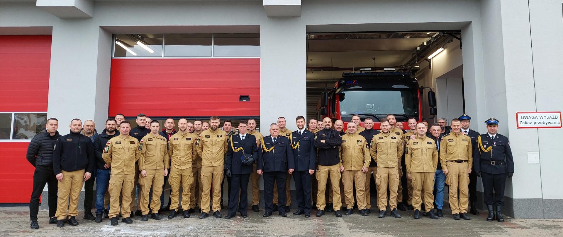 Grupowe zdjęcie strażaków i funkcjonariuszy w mundurach przed budynkiem strażnicy z czerwonymi bramami; w tle widać wóz strażacki w garażu