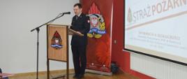 Narada roczna. Świetlica Komendy Powiatowej PSP w Rawiczu. Przy mównicy, na tle ścianki promocyjnej z logo PSP przemawia komendant powiatowy. Strażak ubrany jest w mundur wyjściowy. Z prawej strony znajduje się ekran, na którym wyświetlana jest prezentacja multimedialna. 