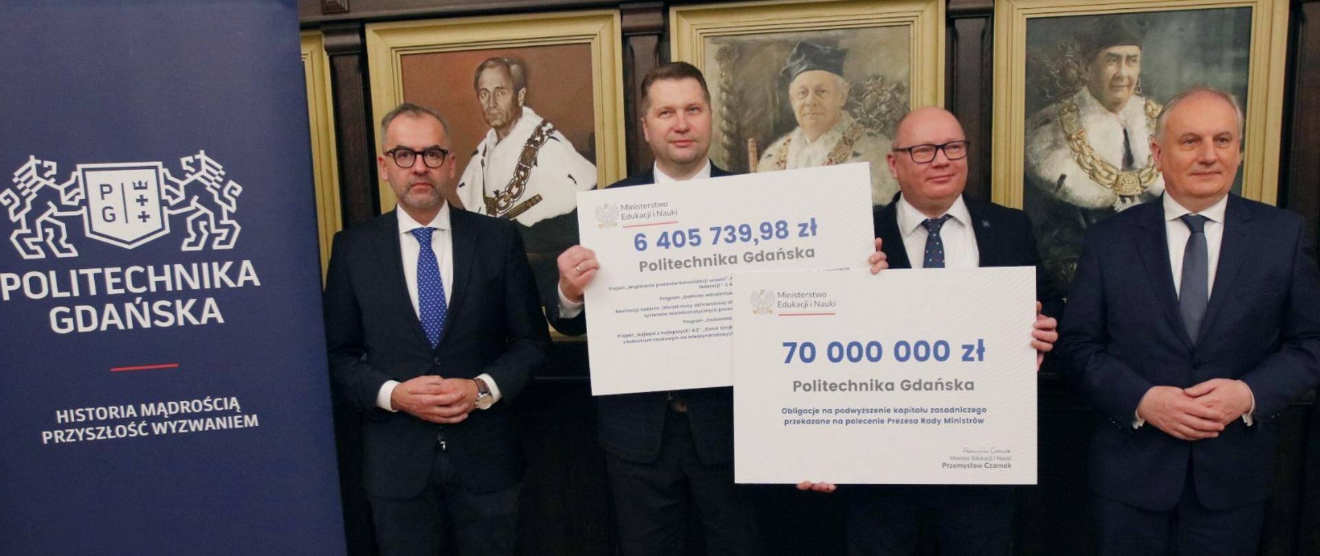 Minister Czarnek i trzech mężczyzn w garniturach stoją na tle ściany, na której wiszą portrety w złoconych ramach. Minister i jeden mężczyzna trzymają wielkie symboliczne czeki z napisami 6 405 739,98 zł i 70 000 000 zł.