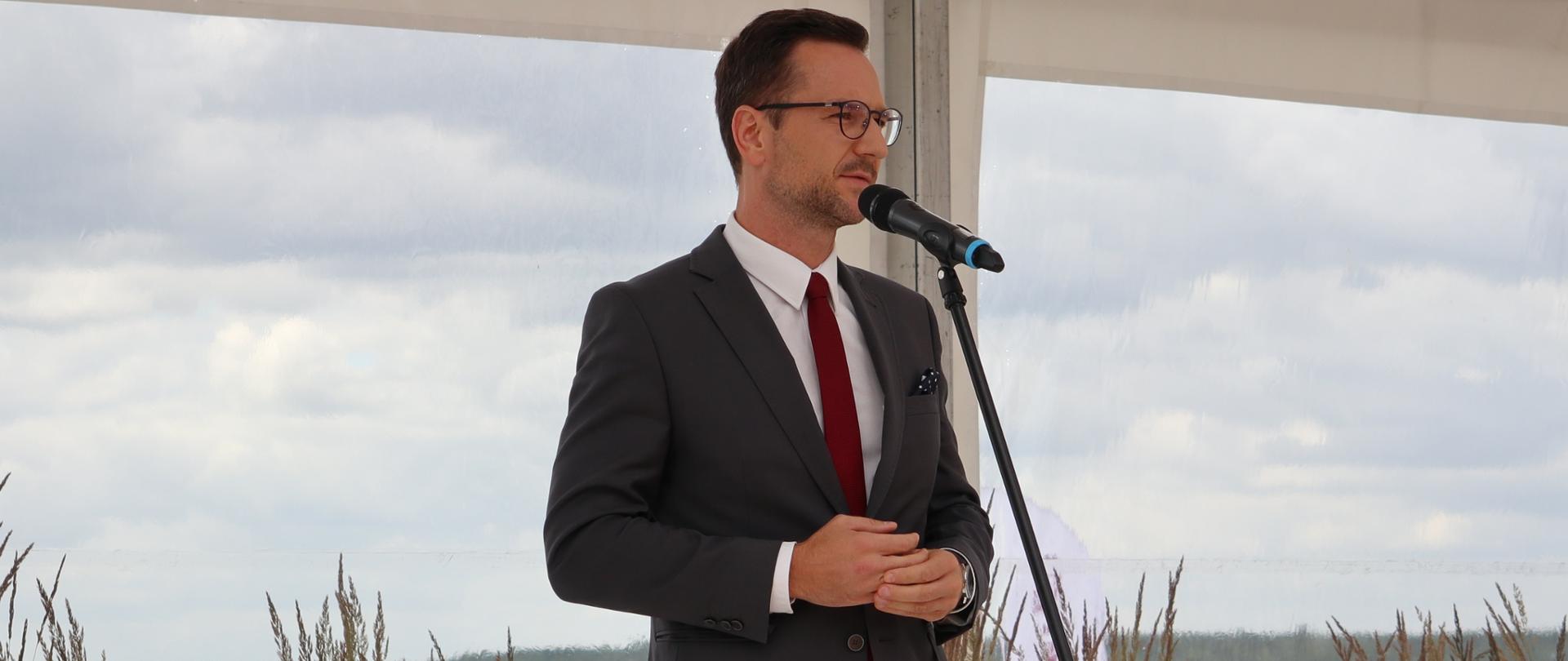 W plenerze, na tle nizinnego krajobrazu przed mikrofonem, stoi wiceminister Waldemar Buda