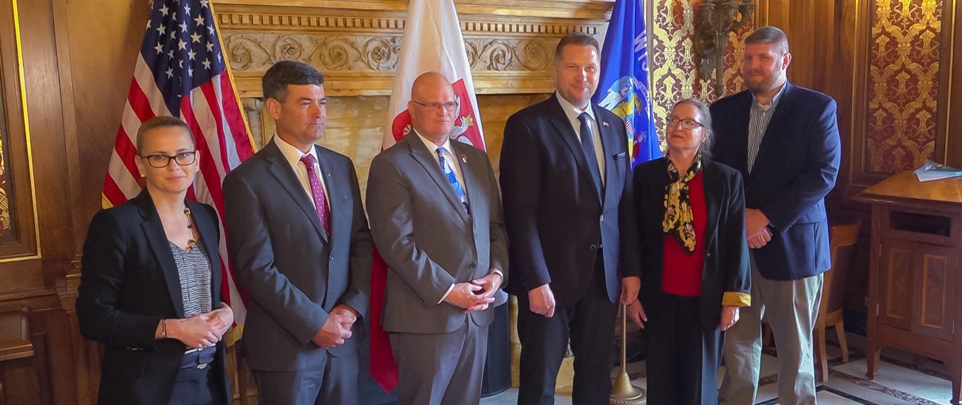 Grupa osob stoi na tle drewnianej, rzeźbionej ściany. Są wśród nih dwie kobiety i trzech mężczyzn. Po środku Minister Przemysław Czarnek. W tle flagi: amerykańska, polska i stanu Wisconsin