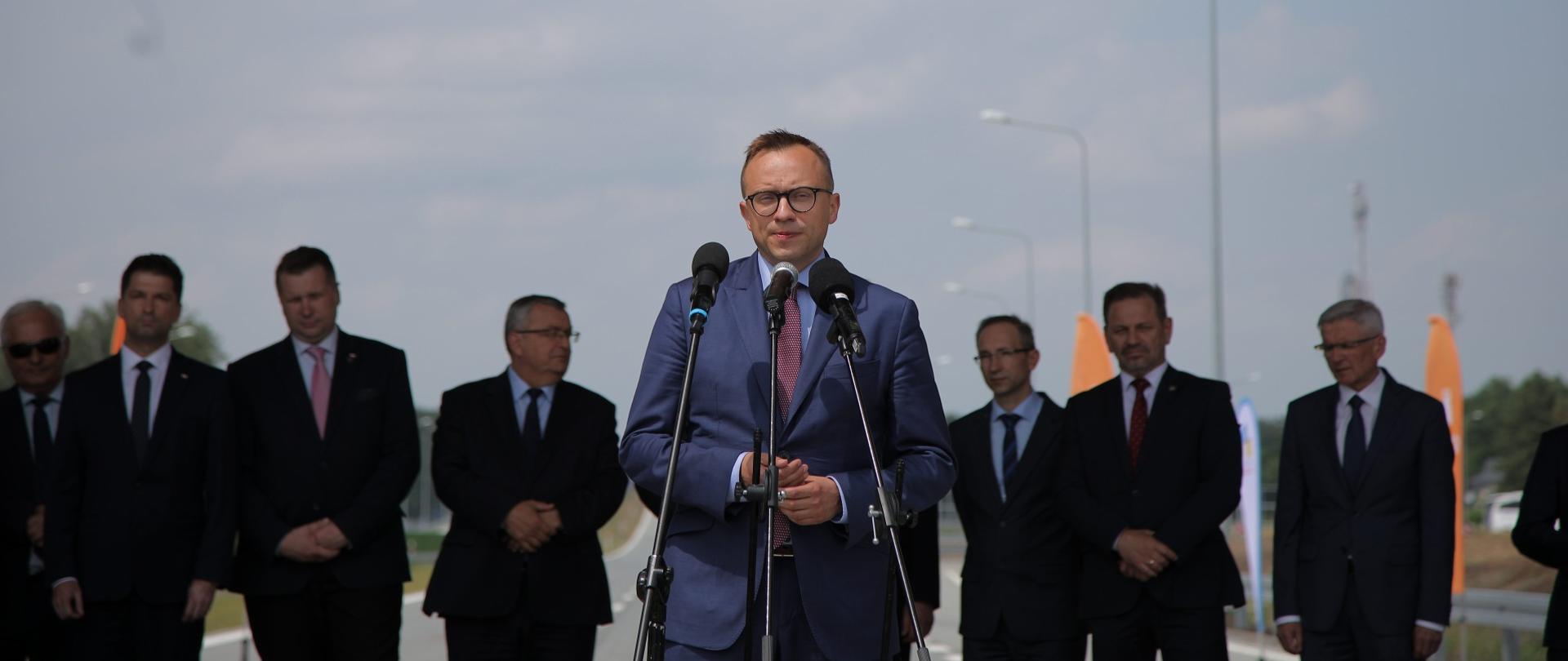 Minister Soboń stoi przy trzech mikrofonach. Za nimi stoi grupa mężczyzn w garniturach. 