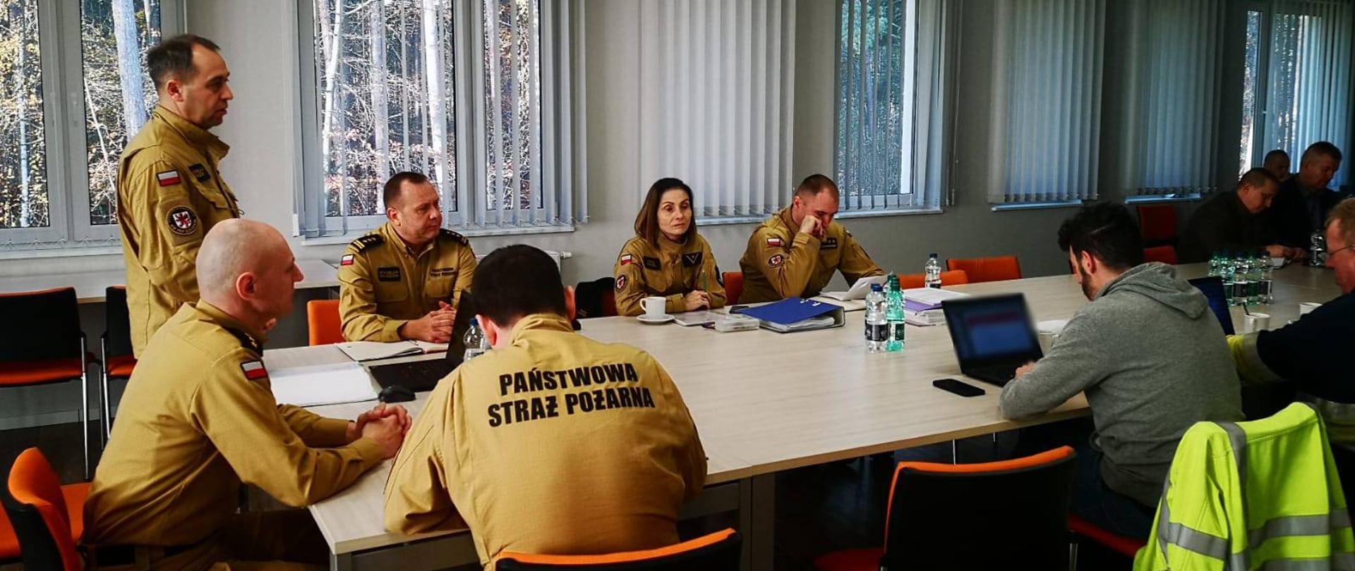 Zdjęcie przedstawia funkcjonariuszy Państwowej Straży Pożarnej oraz przedstawicieli innych służb w trakcie omawiania ćwiczeń. Wszyscy siedzą przy dużym stole. Widać stojącego Zachodniopomorskiego Komendanta PSP witającego wszystkich zgromadzonych oraz rozpoczynającego ćwiczenia.