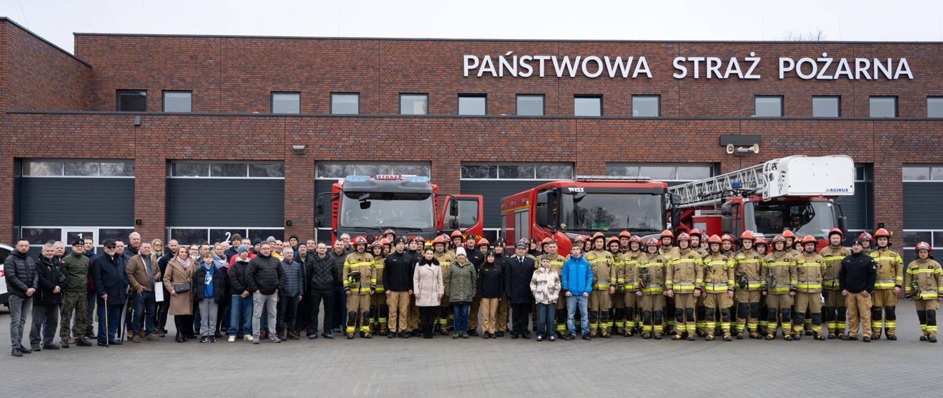 Grupa strażaków w umundurowaniu bojowym oraz funkcjonariuszy i gości cywilnych stoi przed budynkiem z napisem „Państwowa Straż Pożarna”. W tle widoczne są dwa czerwone wozy strażackie zaparkowane przed garażami jednostki.