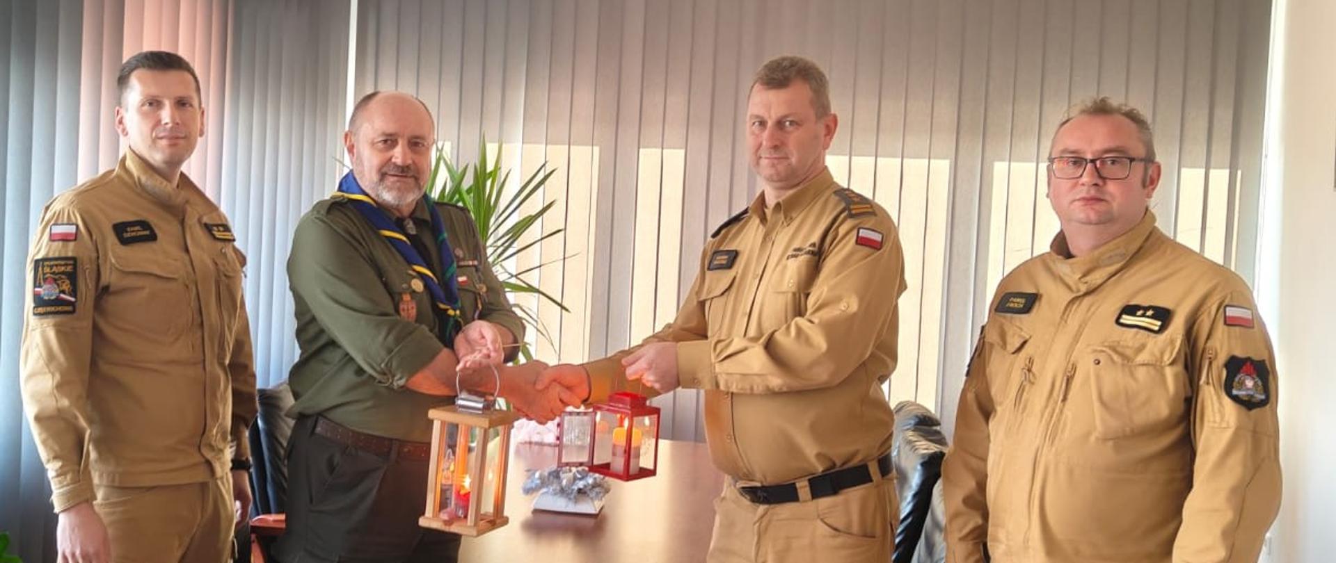 Czterech mężczyzn stoi w biurze obok drewnianego stołu. Mężczyzna po lewej, ubrany w zielony mundur harcerski z żółto-niebieską chustą, trzyma drewniany lampion z zapalonymi świecami. Uściska dłoń mężczyzny po prawej, który jest ubrany w jasnobrązowy mundur służbowy. Pozostali dwaj mężczyźni w identycznych jasnobrązowych mundurach stoją obok, po obu stronach. W tle widać duże okno z jasnymi żaluzjami i roślinę doniczkową.