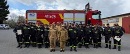 Zdjęcie przedstawia dużą grupę strażaków ustawionych do wspólnej fotografii na utwardzonym placu. Większość osób ma ciemne mundury z żółtymi elementami odblaskowymi oraz czapki, a kilka osób stojących z przodu nosi jasne, piaskowe stroje służbowe. Niektóre osoby trzymają dokumenty lub certyfikaty. Za grupą stoi duży czerwony samochód strażacki z numerem „441[26]” widocznym na boku. W tle znajdują się drzewa, zaparkowane auta, budynek oraz zachmurzone niebo.