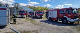 Kilka samochodów pożarniczych ustawionych na terenie ćwiczeń. Widoczne są rozwinięte linie wężowe oraz strażacy przygotowujący elementy zaopatrzenia wodnego.