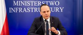 Na zdjęciu widać mężczyznę ubranego w ciemny garnitur, białą koszulę i granatowy krawat. Mężczyzna siedzi przy stole, obok niego jest mikrofon. W jego tle jest granatowa ściana z białym napisem Ministerstwo Infrastruktury.